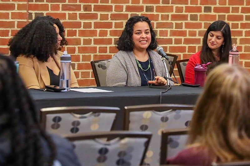Three women sitting at a panel table with a microphone in front of a brick wall.
