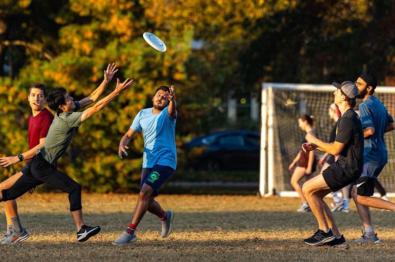 Students play frisbee on a grassy field at sunset with autumn trees in the background.