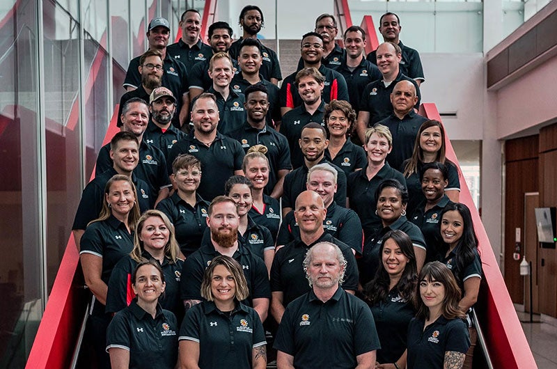 A large group of RecWell staff members in matching black polo shirts posing together on a red staircase.