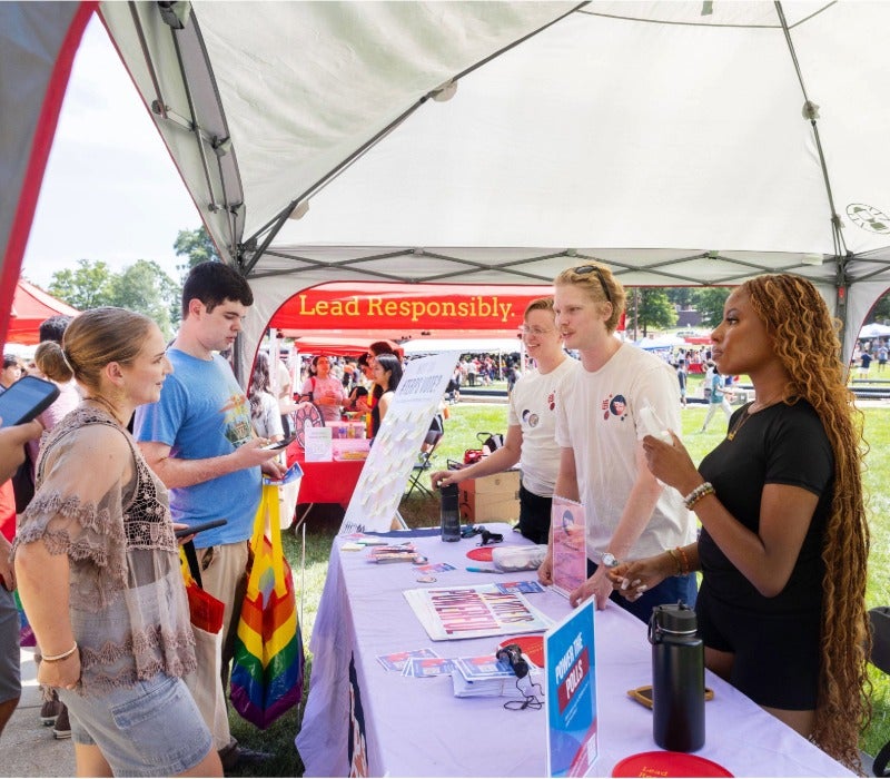 Student leaders chat with visitors at an information table under a tent at an outdoor involvement fair.