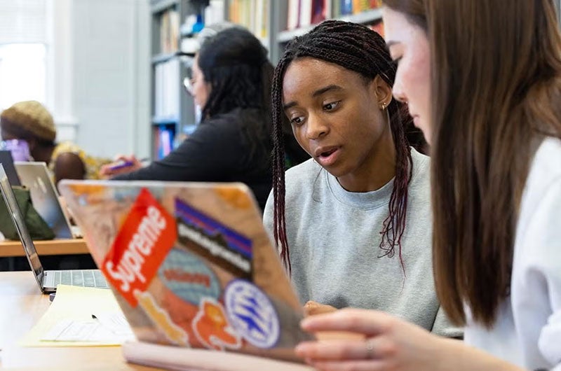 Two students collaborating and looking at a laptop with stickers in a campus library.