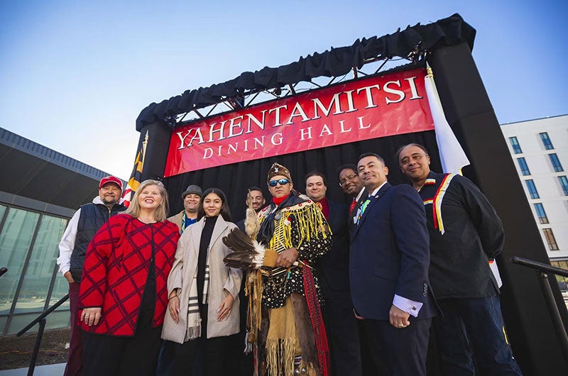 Group of people in regalia and suits stand by a "Yahentamitsi" sign during an outdoor ceremony.