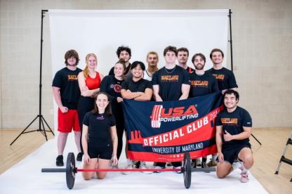 A mixed-gender club sports group poses with their official team banner and a barbell.