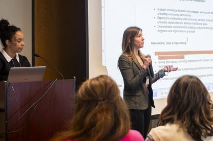 A woman in a gray blazer speaks into a microphone next to a presentation slide on a screen.