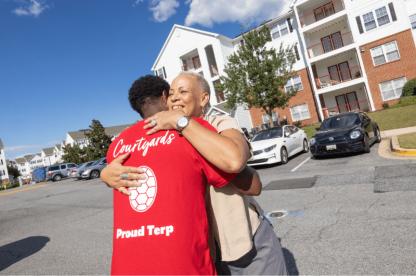 A smiling woman hugs a student wearing a red "Proud Terp" shirt in front of a campus apartment building.