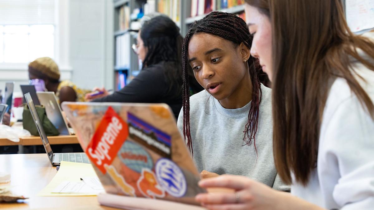 Two students sit at their laptops and are talking with each other 