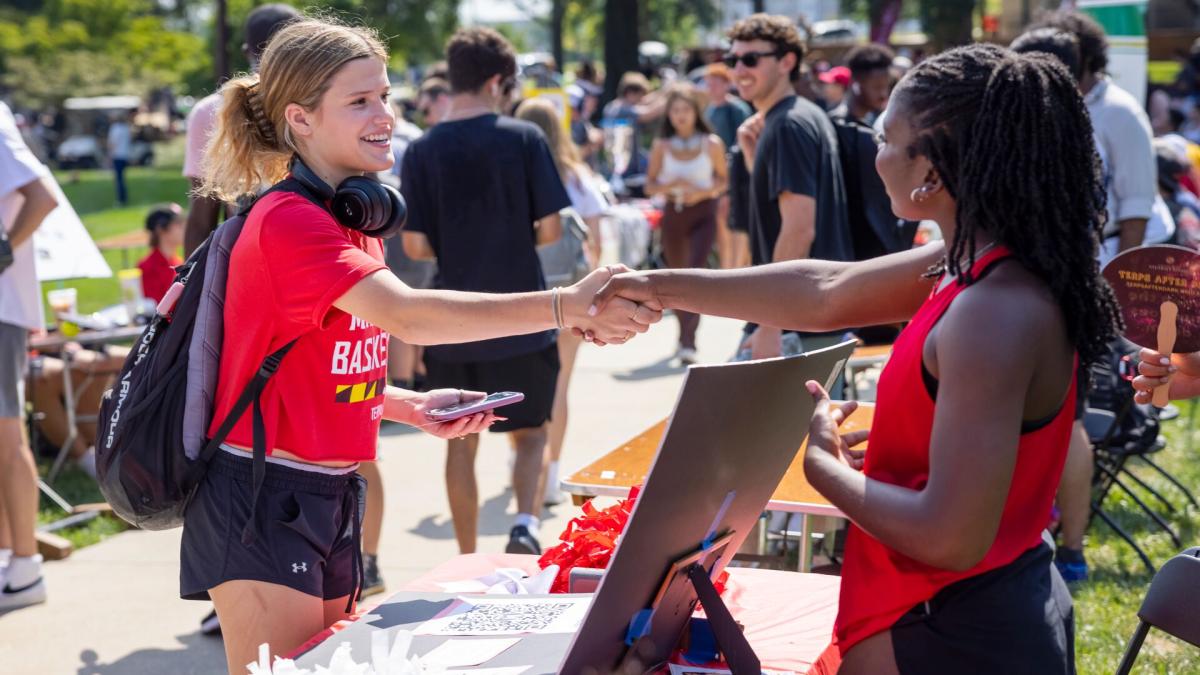 Two students at an outdoor fair shake hands across a table