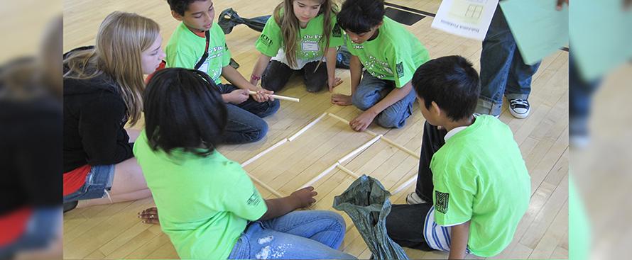 A circle of children collaborating with one another, using wooden dowels to create the shape of a window on the wooden floor