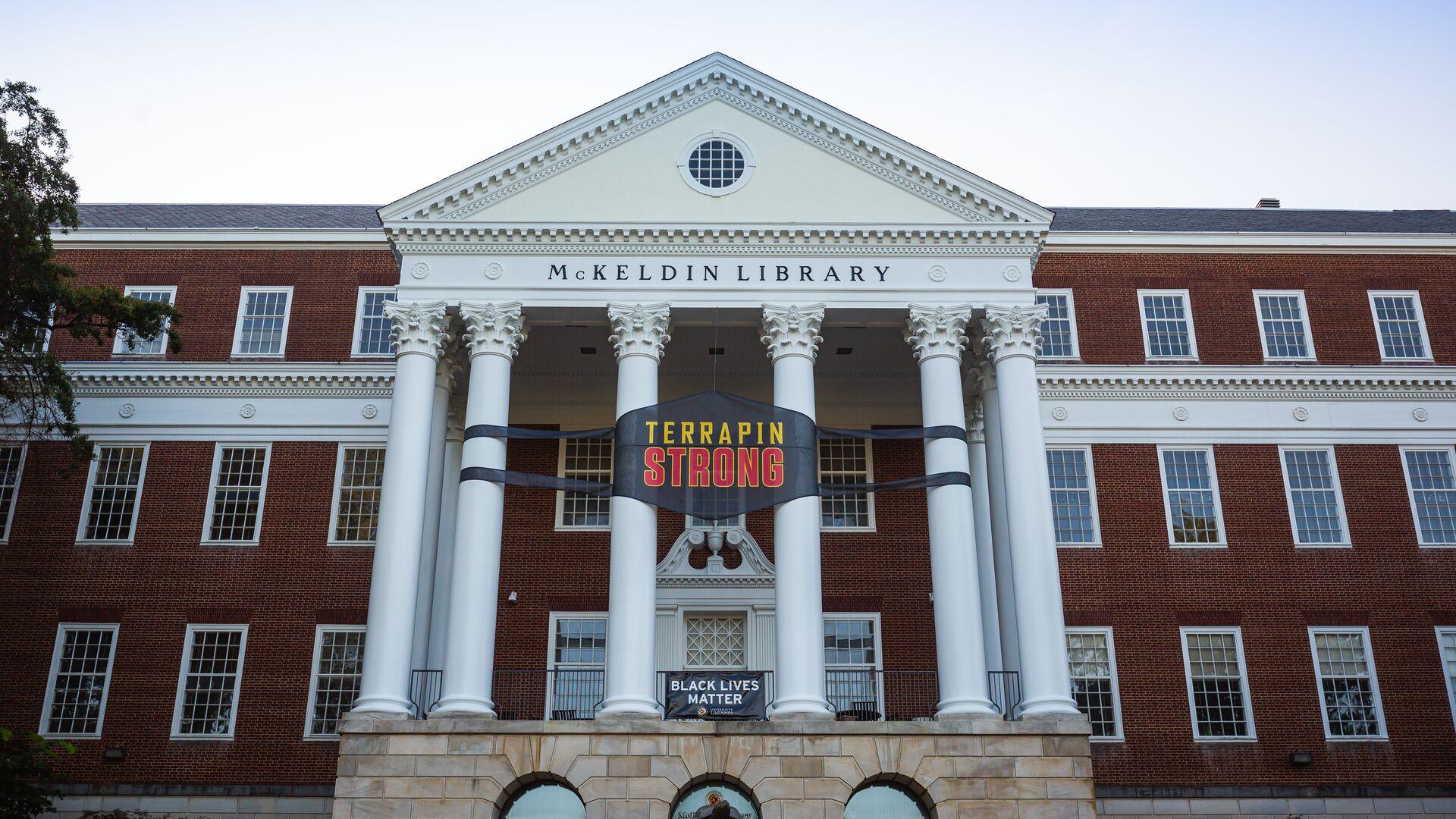 Terrapin Strong "mask" banner on the front exterior of McKeldin Library at dusk; Testudo statue in the foreground.
