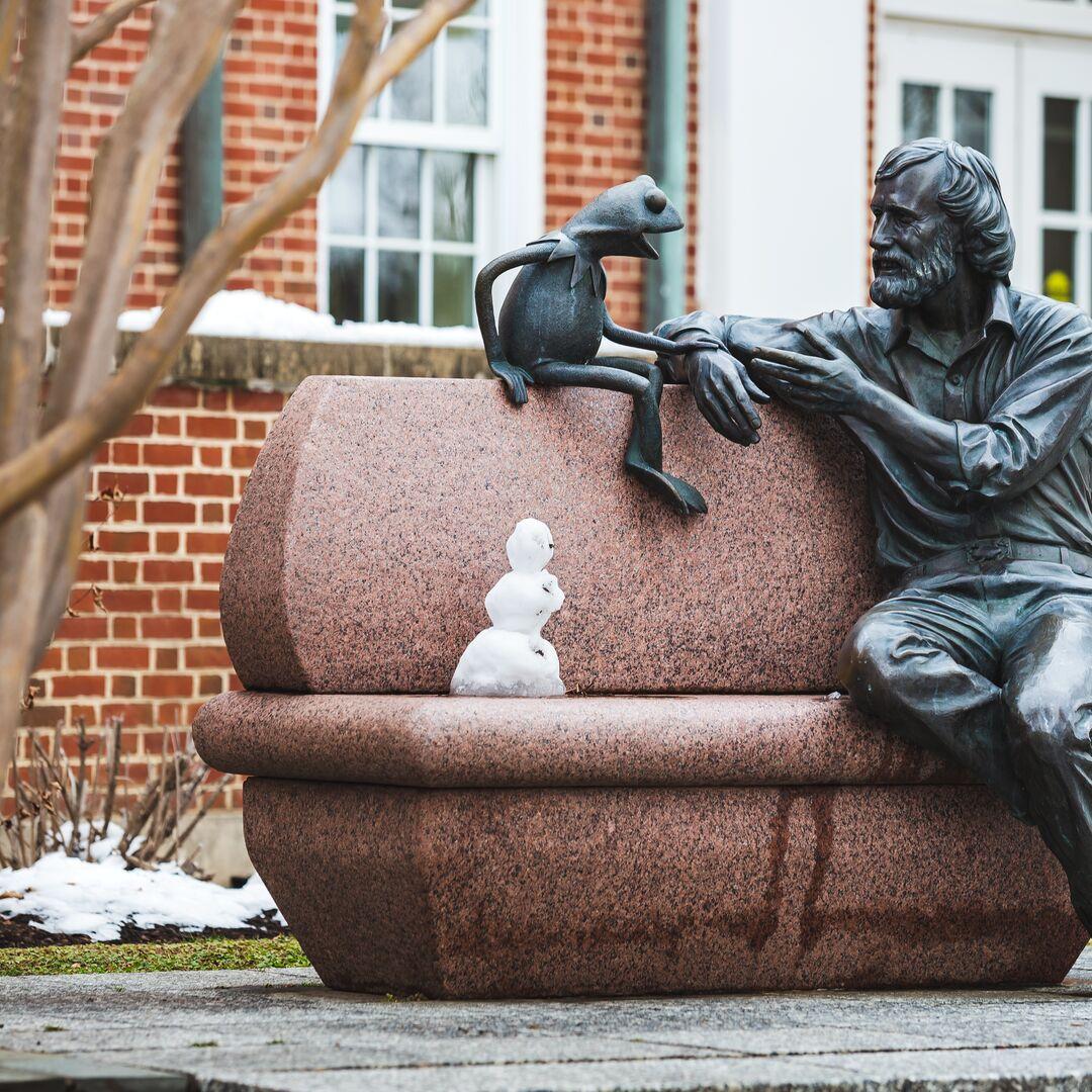 Kermit and Jim Henson statue in winter. A little snowman sits on the bench with them.