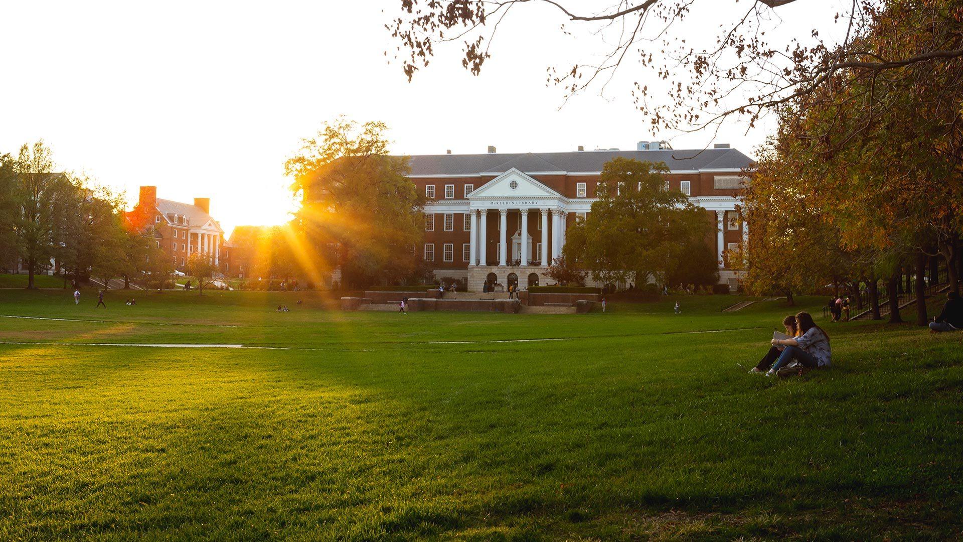 The sun sets next to McKeldin Library in the background while two students sit on the grass in the foreground