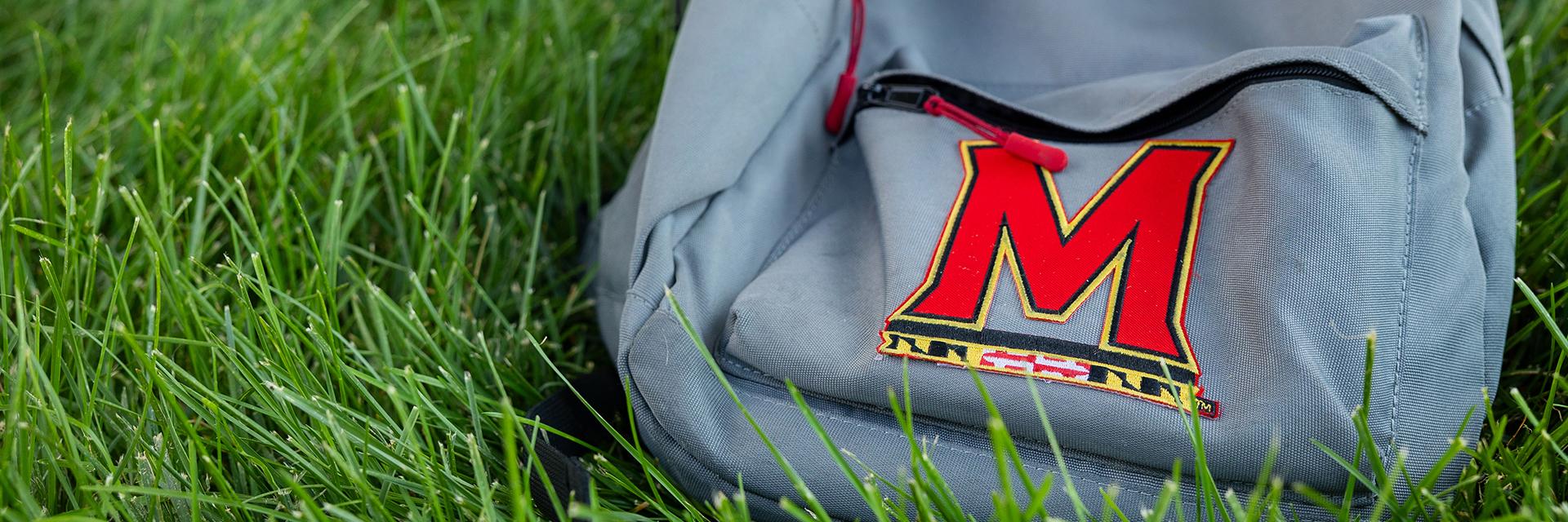 Grey backpack laying in the grass with the red letter M logo
