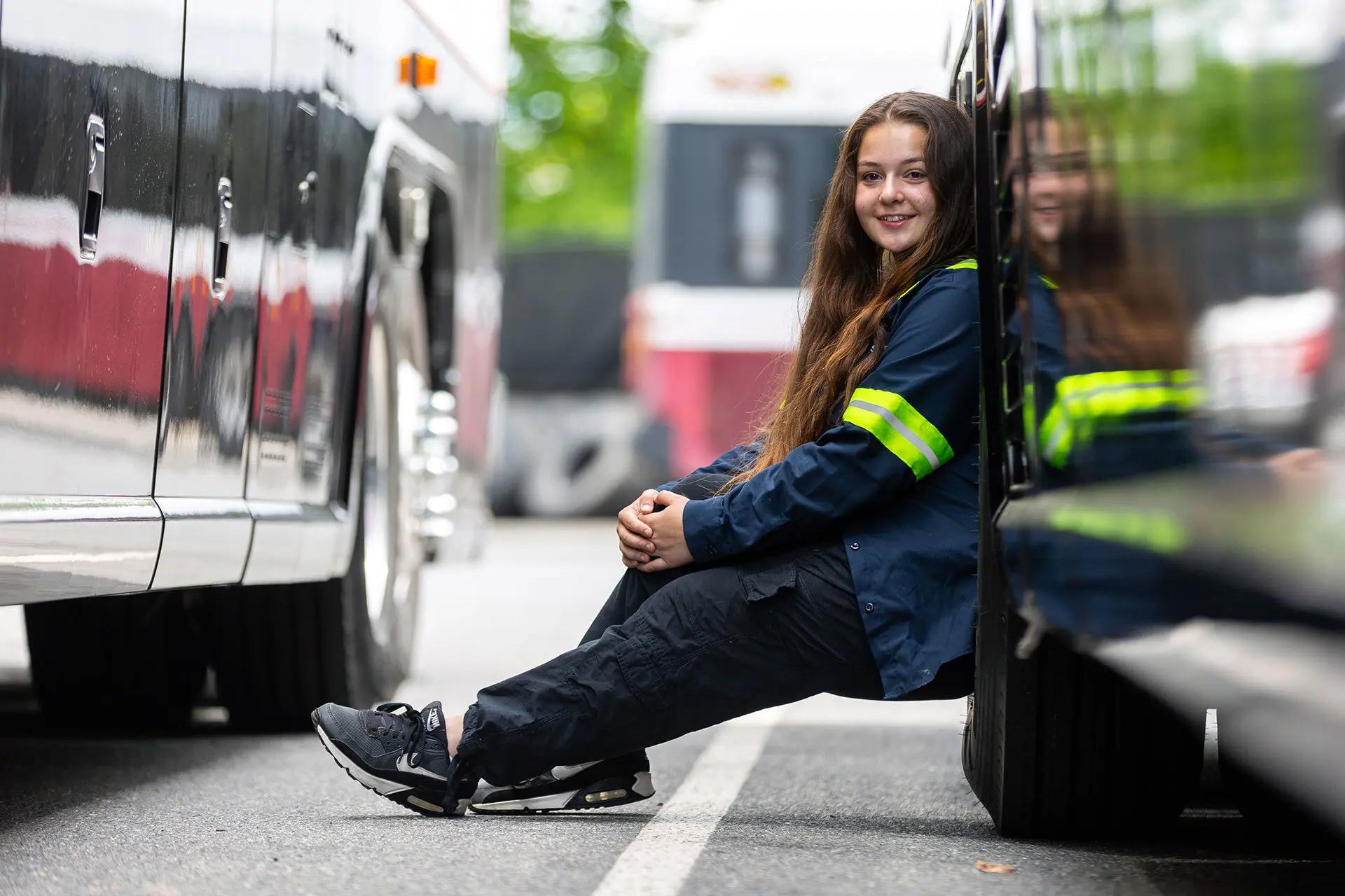 Katie is wearing a work coat with neon yellow striping on the arm, leaning back against the side of a bus, turning toward the camera with a smile.