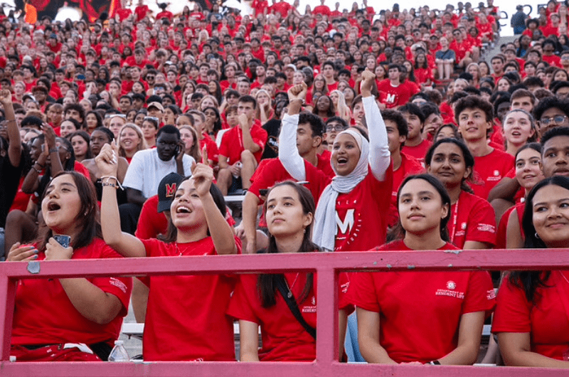 A large crowd of students in red Maryland shirts cheers enthusiastically from the stadium bleachers.