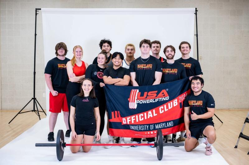 A mixed-gender club sports group poses with their official team banner and a barbell.