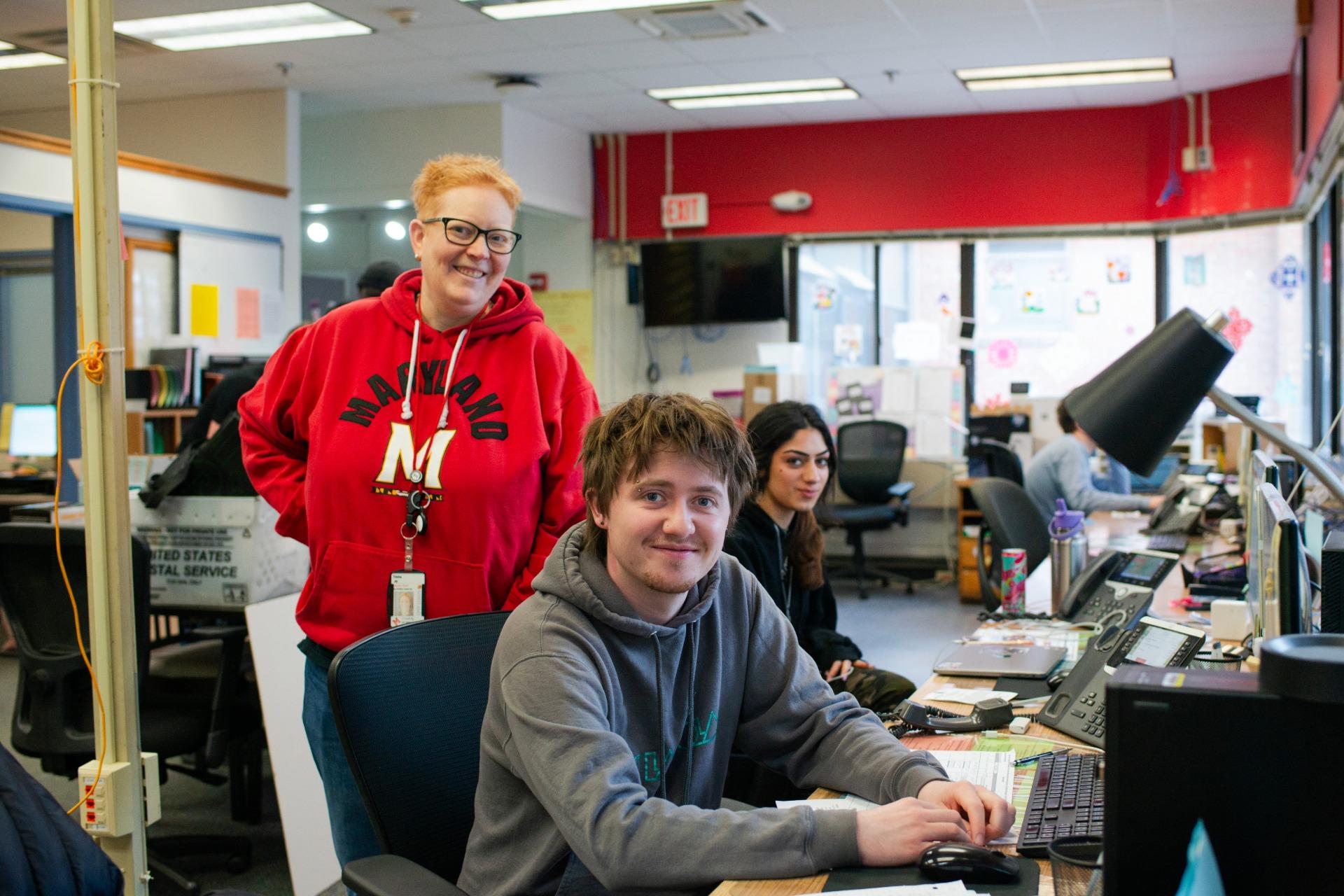 Two students and a staff member smiling in the Residential Facilities Service Center workspace with desks and computers.