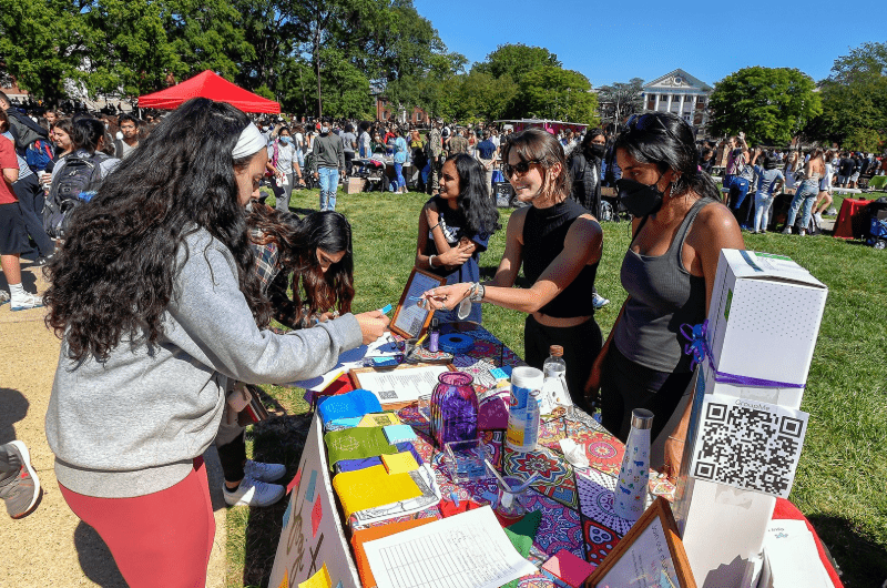 Students interact at a colorful information table during a busy First Look Fair on a sunny lawn.