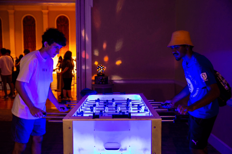Two students play foosball on a glowing LED table during a late-night event.