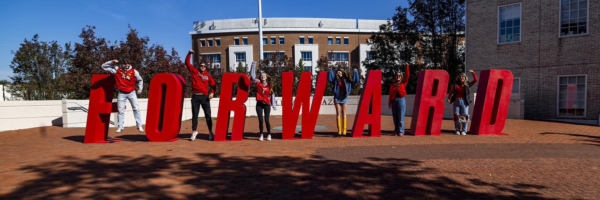 Seven students jump and pose around large red letters spelling "FORWARD" on a brick plaza.