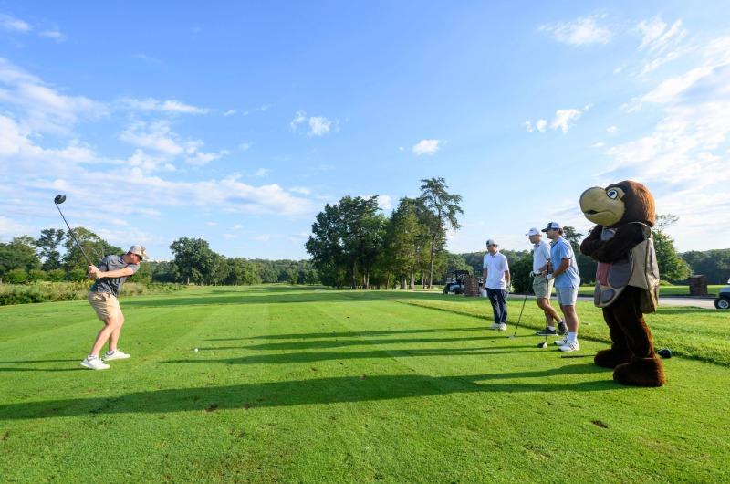 A student swings a golf club on a green while others and Testudo watch.