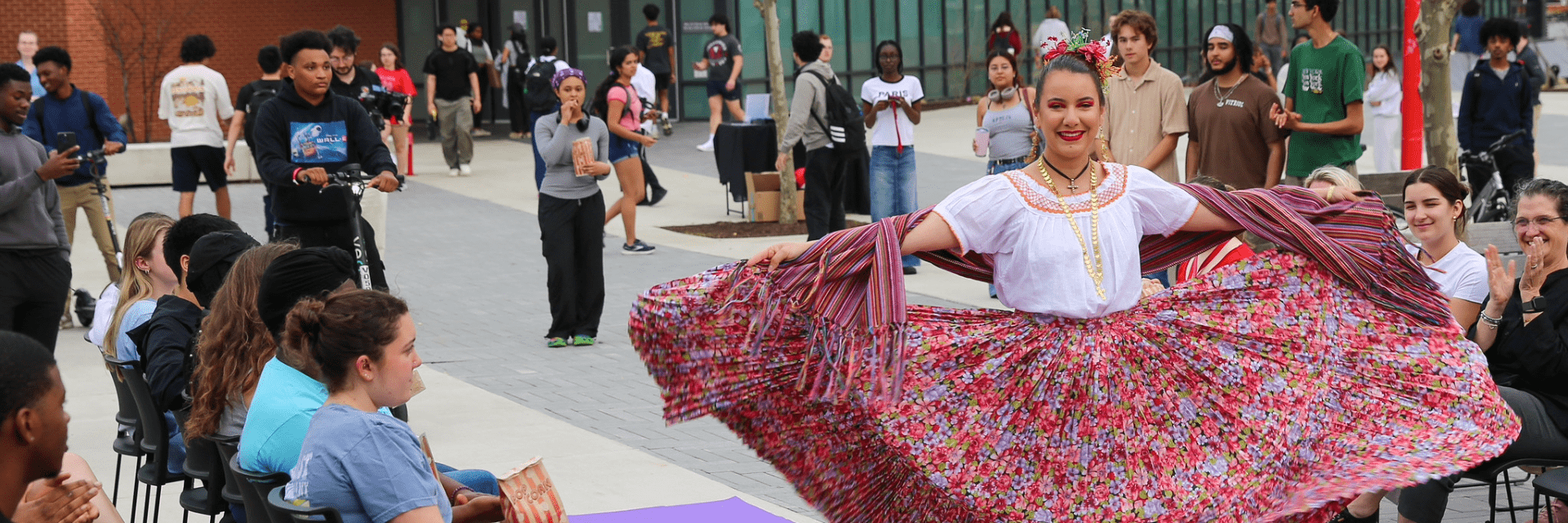 A woman in a vibrant, floral traditional dress walks down a runway for an audience on Heritage plaza.