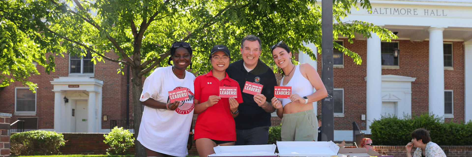 Four people smile and hold "We Are Looking For Leaders!" cards in front of Baltimore Hall at a block party.