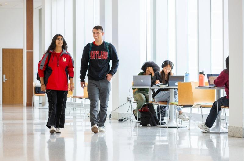 Two students in Maryland apparel walking and talking in a bright, modern hallway with study areas.
