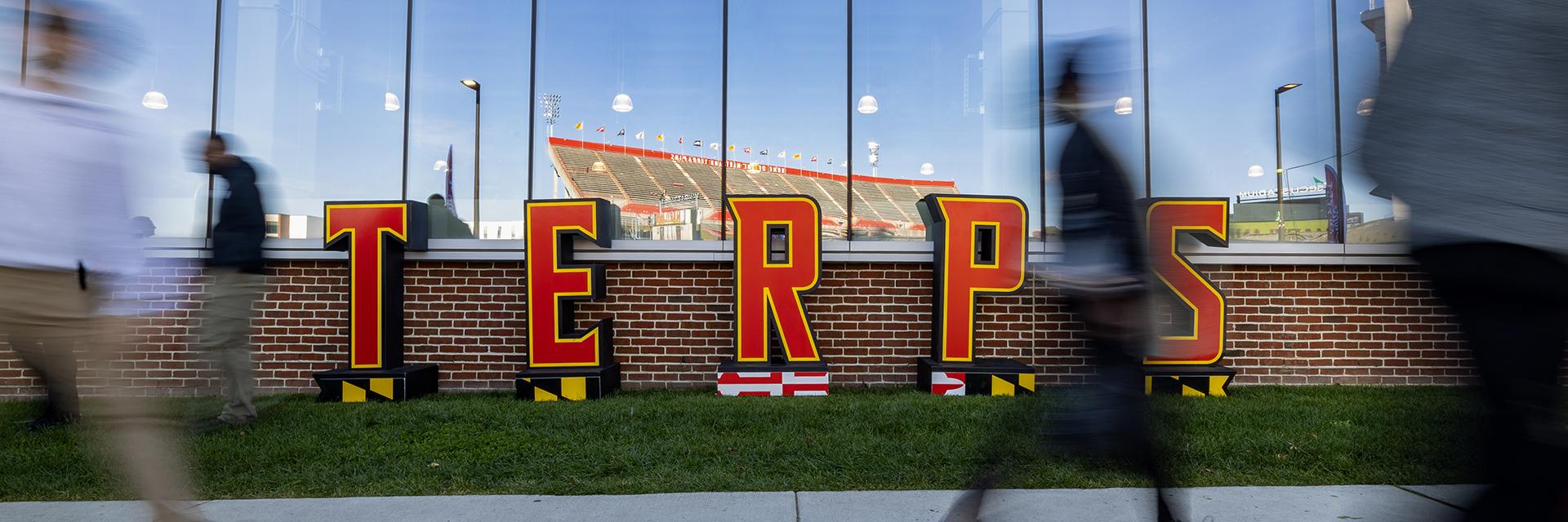 Large red "TERPS" letters in front of a brick wall and a glass building reflecting a stadium.