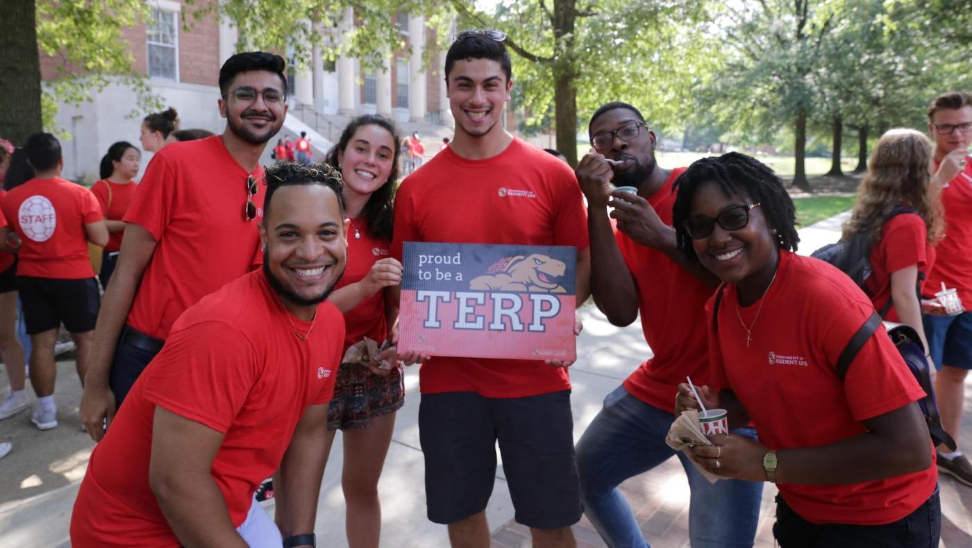Group of Resident Assistants in red shirts smile while holding a "Proud to be a TERP" sign outdoors.