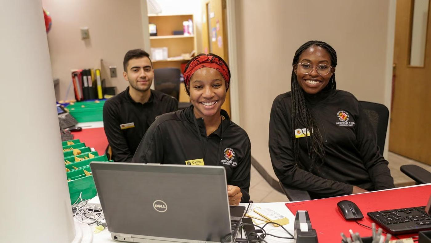 Three student community assistants smile at the Annapolis Hall service desk with a laptop and mail room in the background.
