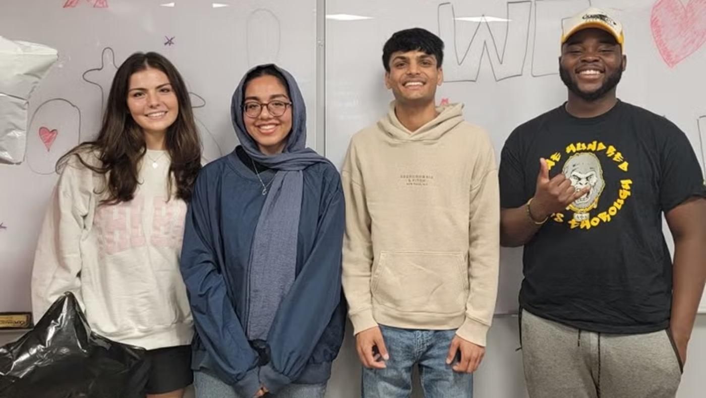 Four smiling student assistants stand side-by-side in front of a whiteboard with drawings.