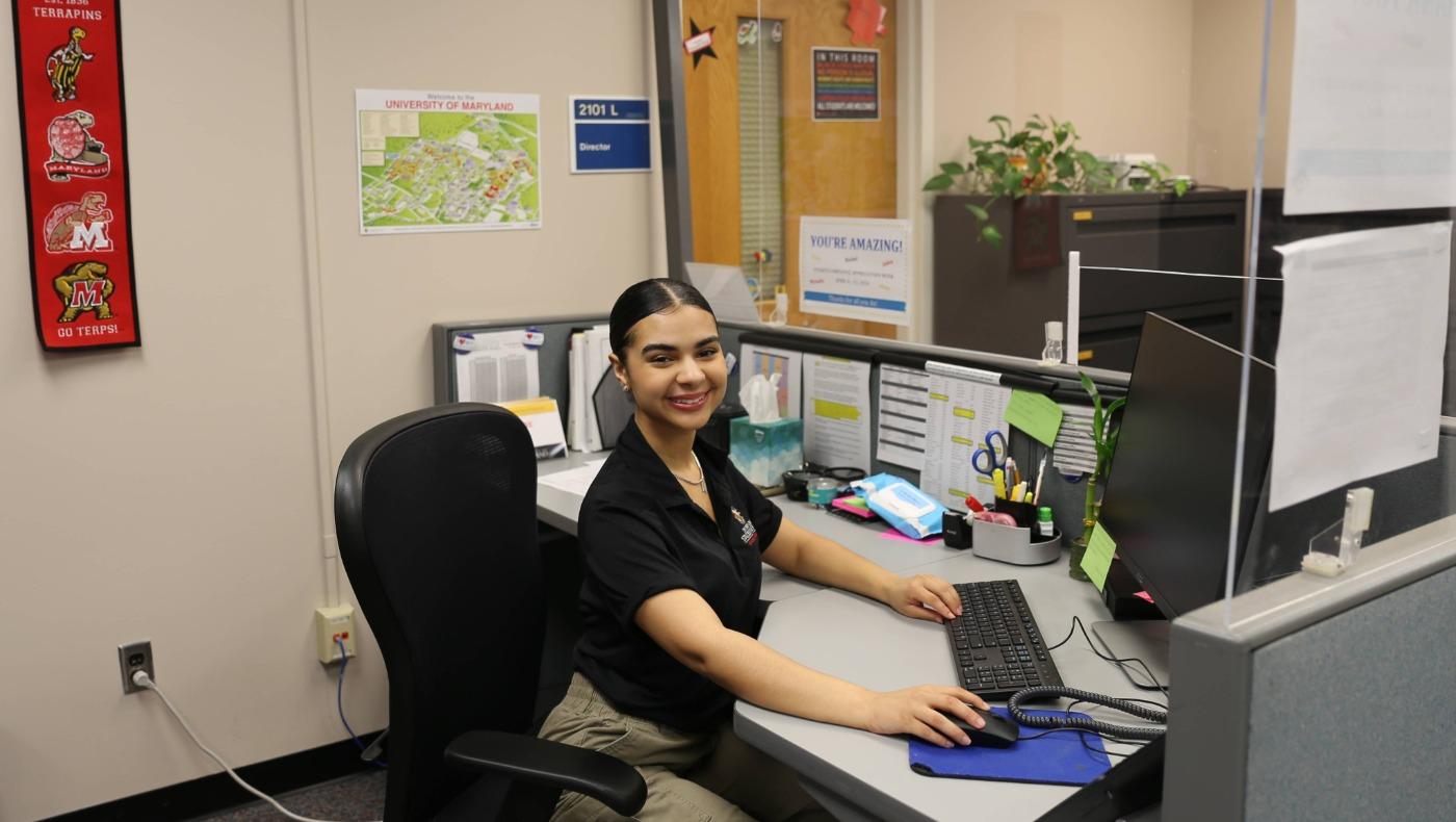 A student office assistant sits at a desk with a computer in the Res Life director's office.