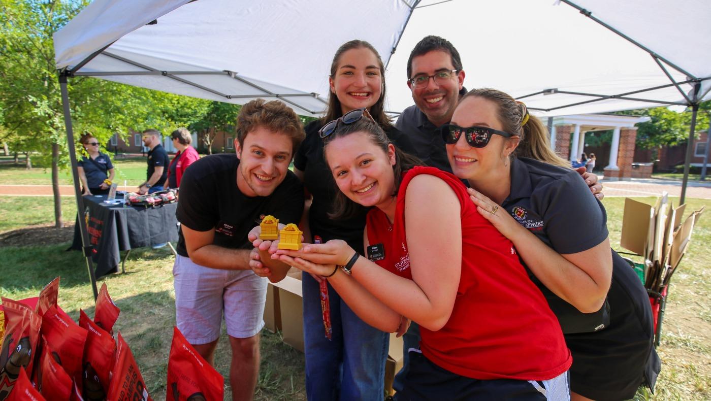 Three program assistants and two staff members posing under a white tent at an outdoor event, holding a 3D printed testudo.