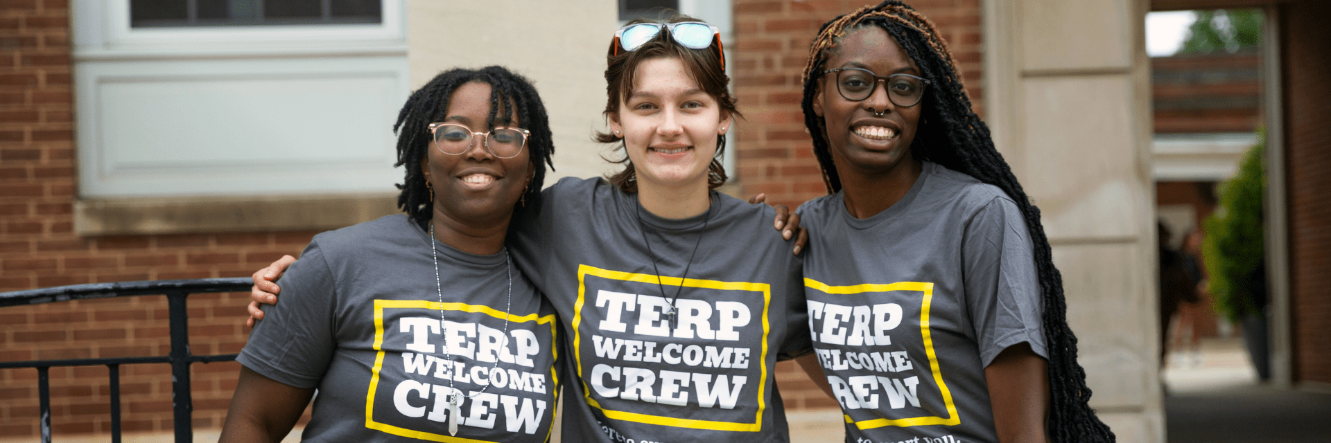 Three students stand smiling with their arms around each other. They are wearing gray "Terp Welcome Crew" t-shirts and are standing in front of a brick building.