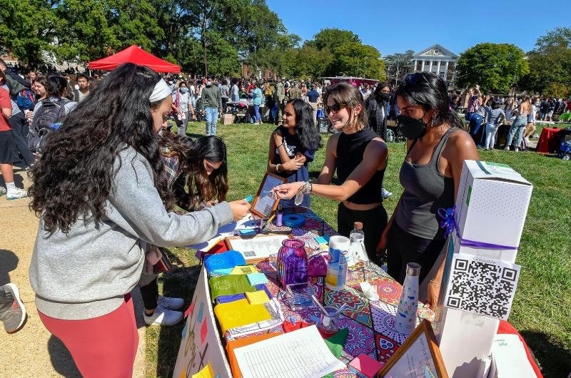 Students interact at a colorful information table during a busy First Look Fair on a sunny lawn.