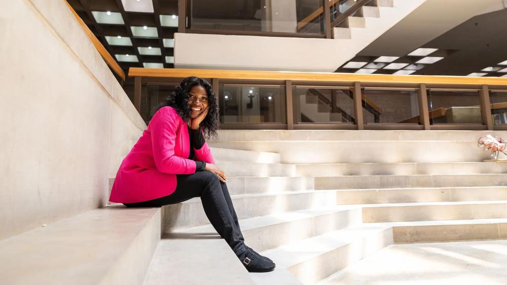 Student Zoh Fopenawoh sitting on steps in black slacks and a pink blazer, leaning forward with her head resting on her hand and a big smile on her face.
